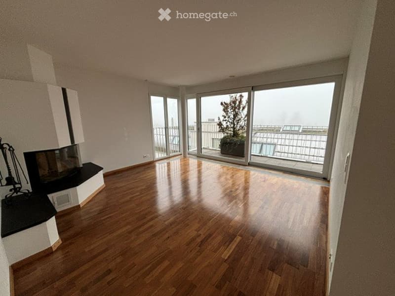 Bright, modern living room with wooden flooring, large glass doors leading to a balcony with a view, and a fireplace on the left side.