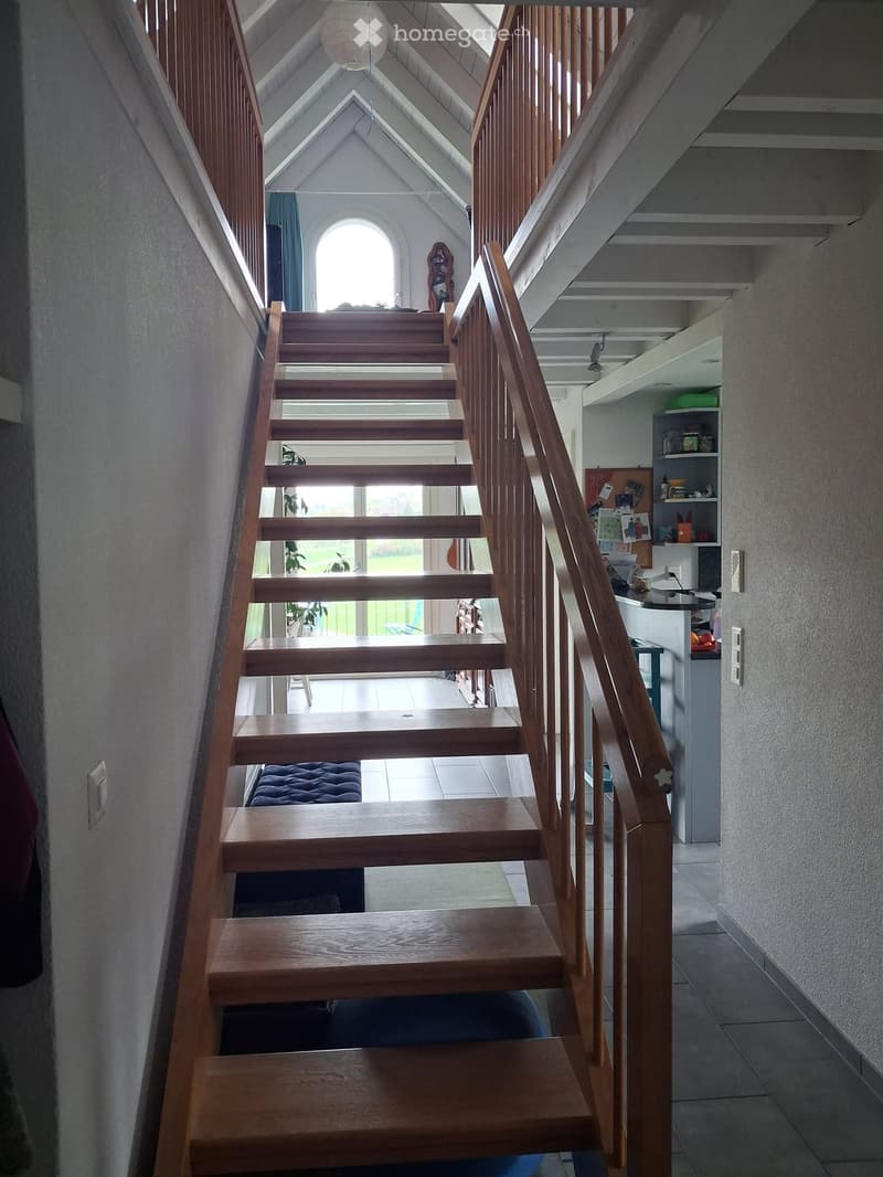 Wooden staircase with a wooden handrail, leading to a bright room with a large arched window and visible kitchen area in the background.