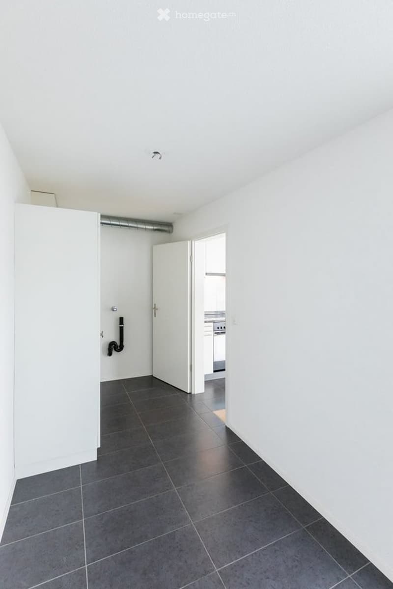Completely empty room with white walls, dark grey tiled floor, and a doorway leading to another room.