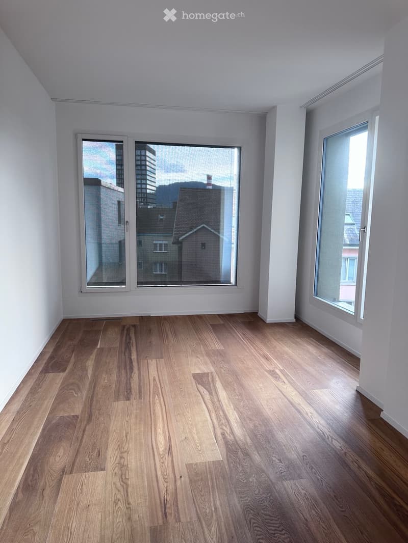 Empty room with wooden flooring and large windows offering a cityscape view.