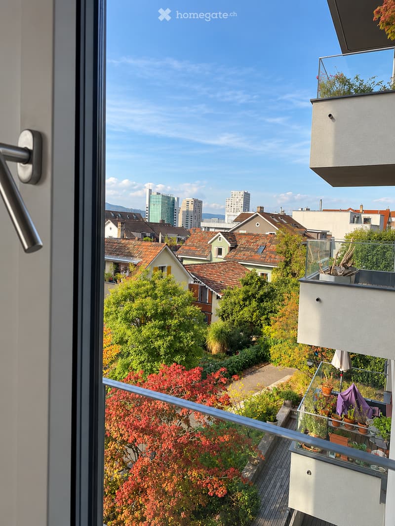 Balcony with glass railing, potted plants, and a view of a cityscape with trees and buildings.