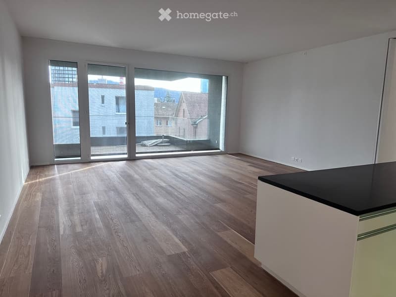 Empty room with wooden flooring, large windows, and a small kitchen counter visible on the right side.