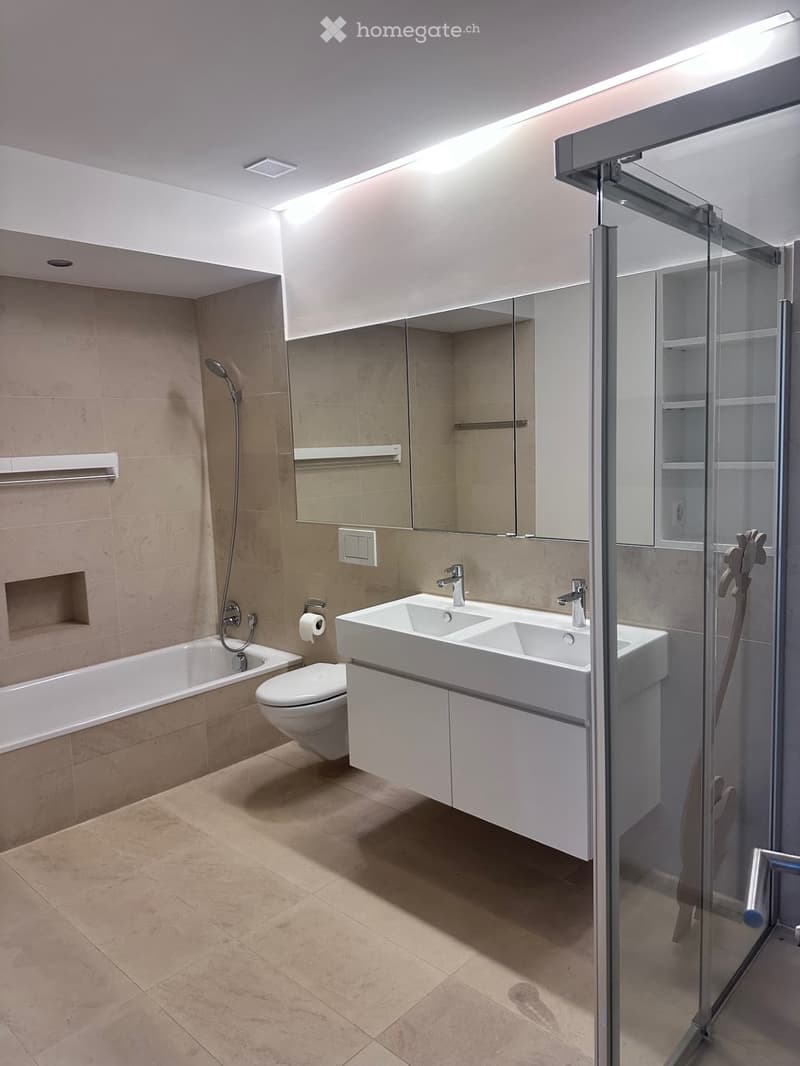 Modern bathroom with beige tiles, a bathtub, toilet, double sink, large mirror, and a glass-enclosed shower area.