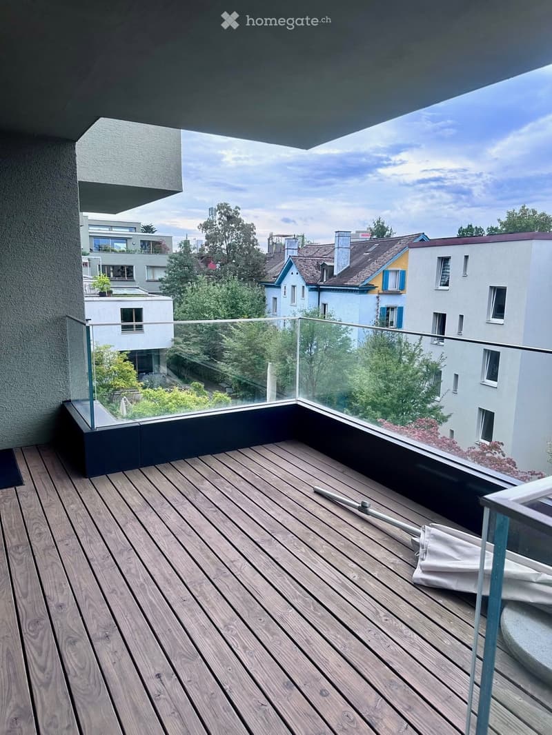 Wooden deck with glass railing, overlooking a cityscape with trees and buildings.