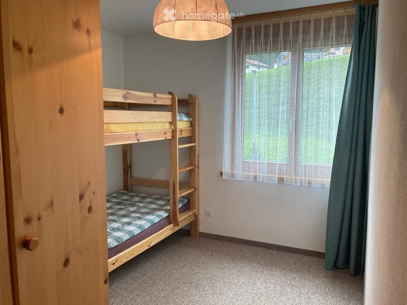 A bedroom with a wooden bunk bed, a window with white curtains, and a power outlet on the wall.