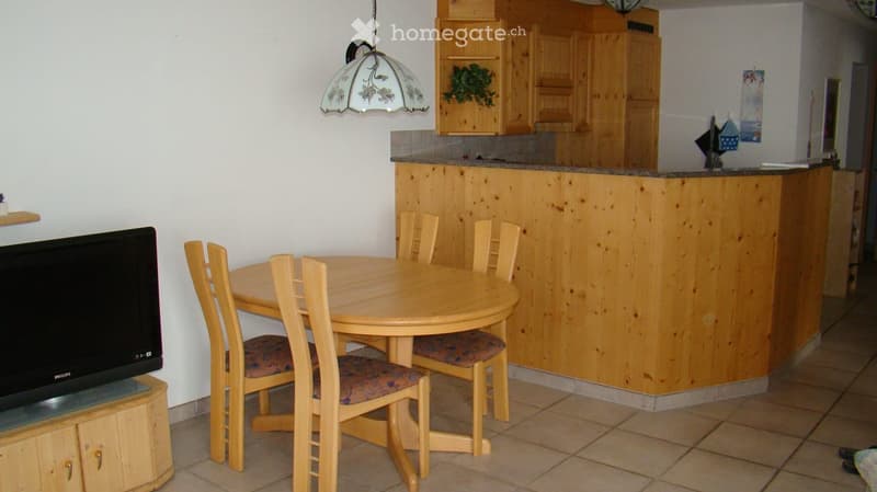 Dining area with a round wooden table and chairs, a flat screen TV on a wooden stand, a wooden counter with a bar stool, wooden cabinets, and a plant on the wall.