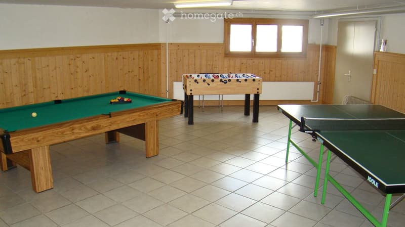 A room with a billiard table, a foosball table, and a ping pong table. The walls are wooden, and there is a window with a radiator below it.