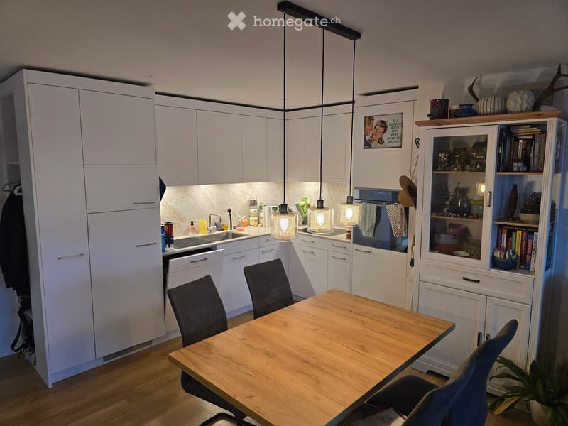 Modern kitchen with white cabinets, a wooden dining table, four chairs, and a sink area with a faucet. There is also a bookshelf with books and decorative items.
