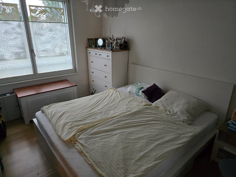 Bedroom with a made bed, nightstand, mirror, drawer unit, and a small table near the window.