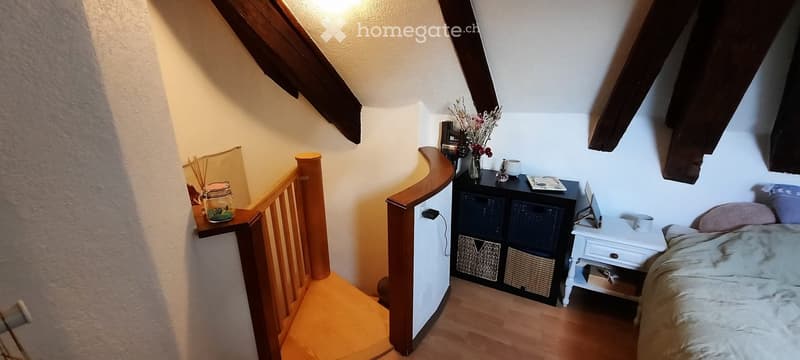 Cozy attic bedroom with wooden slanted ceilings, a bed, nightstand, storage unit, and decorative items including a vase with flowers and a laptop on the nightstand.