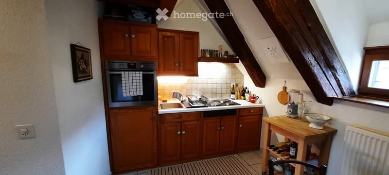 Cozy attic kitchen with wooden cabinets, stainless steel oven, stove, sink, and a small wooden table with various kitchen items.