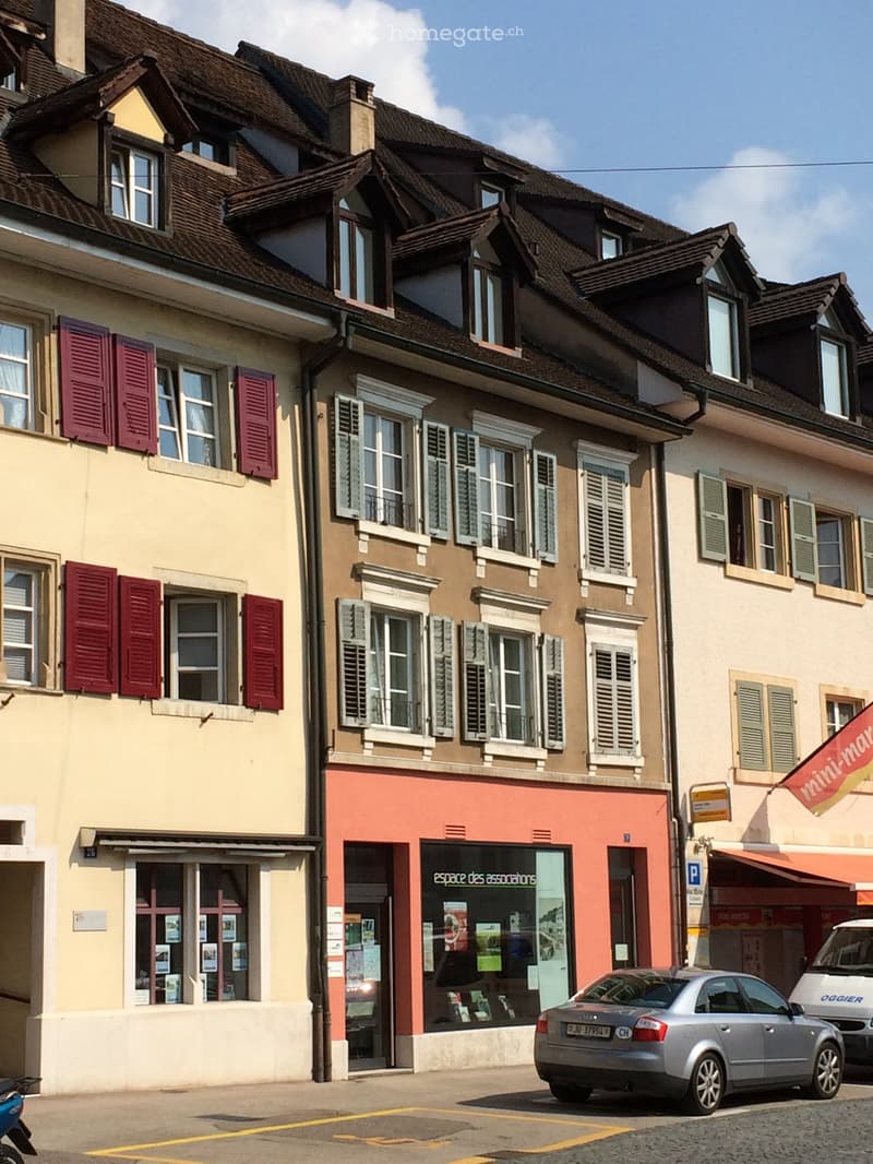 Three-story building with multiple windows, shutters, and a storefront labeled "espace des associations" with posters and flyers visible inside. A silver car is parked in front of the building.