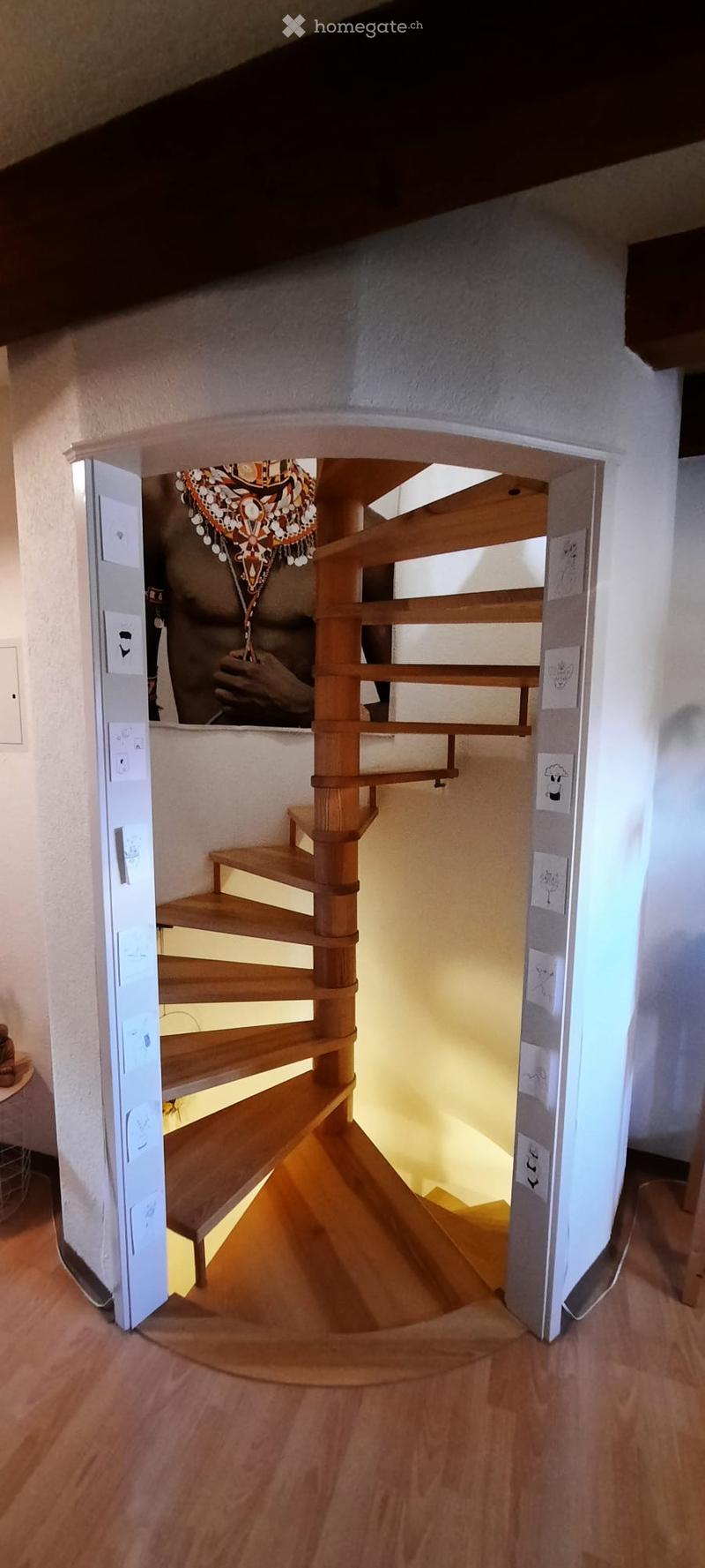Spiral wooden staircase with a handrail, leading upwards, with a framed picture on the wall and a light switch visible on the left side.