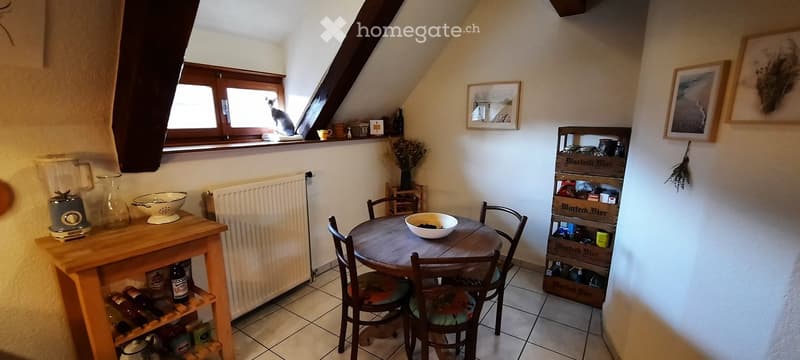 A cozy, well-lit room with a round wooden table and chairs, a small wooden shelf with various items, a blender on a counter, and a cat sitting on a windowsill. The room features framed pictures on the walls and a radiator under the window.