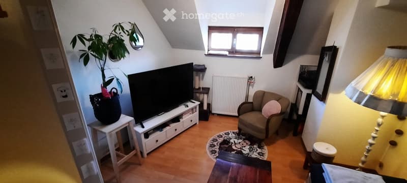 A cozy living room with wooden flooring, a large window, a sofa, a chair, a lamp, a screen, a table, a stool, a plant, and a mirror.