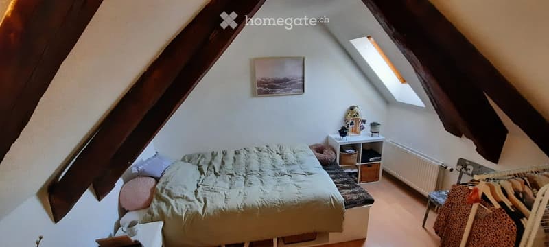 Cozy attic bedroom with slanted wooden beams, a bed with a light green duvet, a small nightstand, a radiator, and a wardrobe with hanging clothes.
