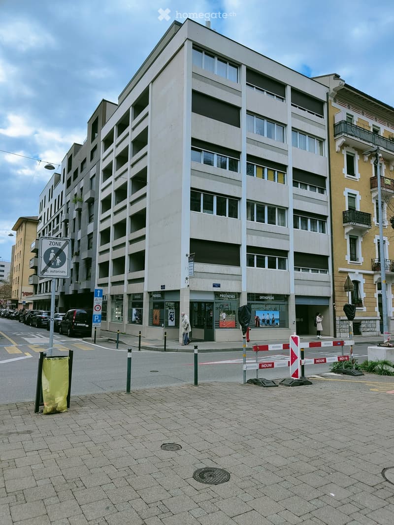 A modern multi-story building with large windows and commercial storefronts at the ground level, located on a paved street with traffic signs and barriers.