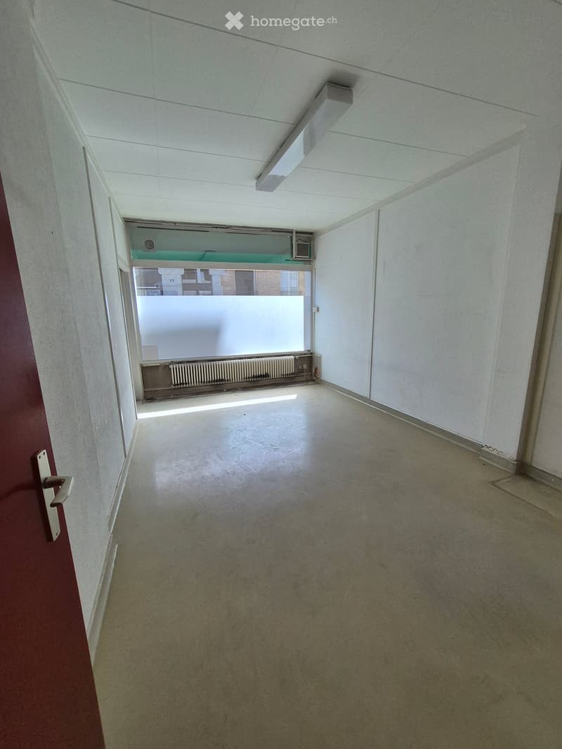 Empty room with bare walls, concrete floor, and a single door. Natural light enters through a window with frosted glass.