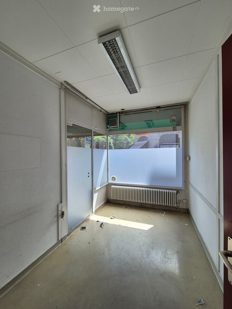Empty hallway with concrete floor, white walls, a radiator, and a large window with blinds partially open, allowing natural light to enter.