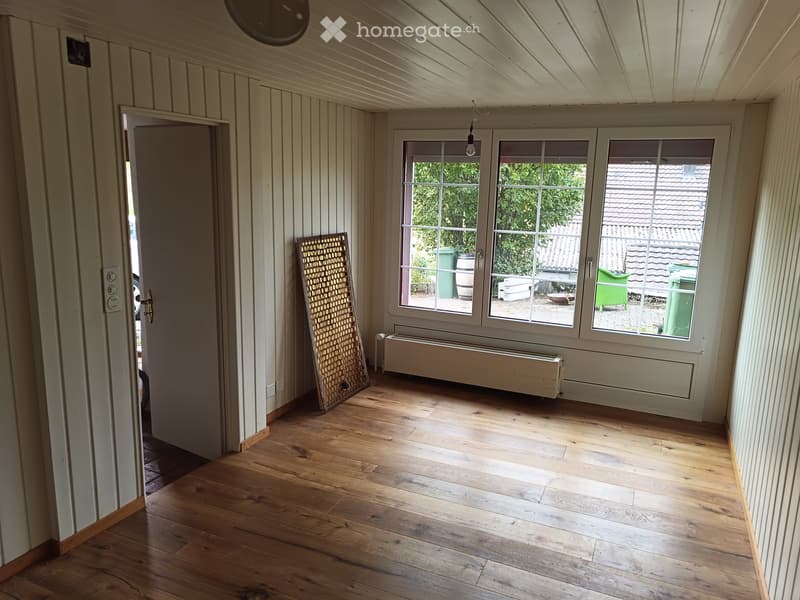 Empty room with wooden flooring, white paneled walls, large windows, and a radiator. A screen is leaning against the wall near the windows.