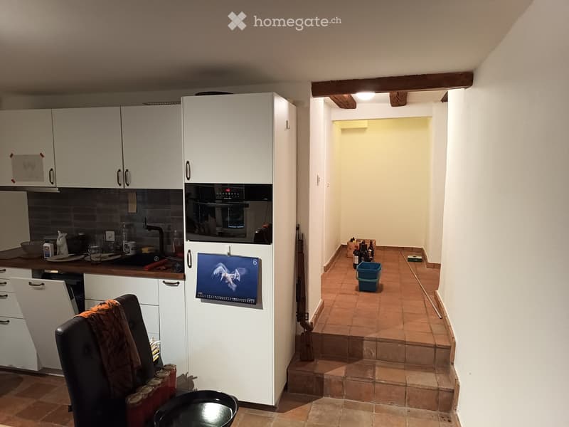 A small kitchen area with white cabinets, a refrigerator, and an oven. A chair with a jacket draped over it is positioned near the counter. A small hallway leads to another room with tiled flooring and a few items, including bottles and a blue bucket.