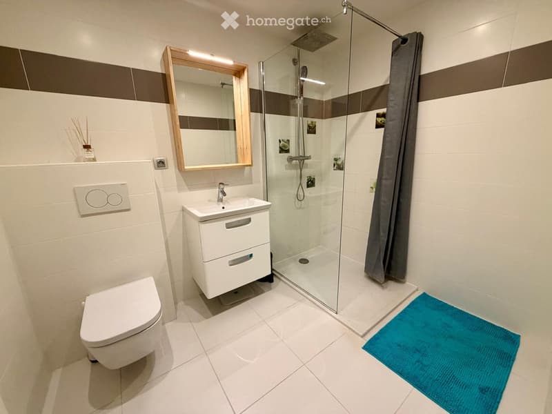 Modern bathroom with white tiles, a toilet, a sink with a cabinet, a glass-enclosed shower, a mirror with wooden frame, and a blue bath mat.