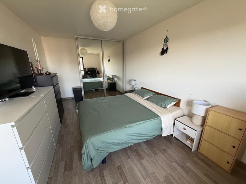 Modern bedroom with a large bed, nightstand, lamp, dresser, and a large mirror reflecting another room.