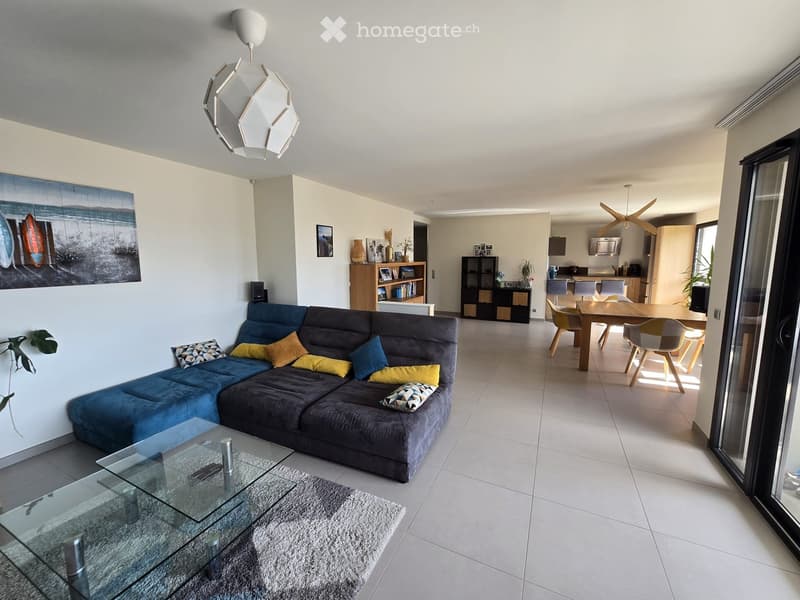 Modern living room with a blue sectional sofa, glass coffee table, dining area with wooden table and chairs, and a kitchen area visible in the background.