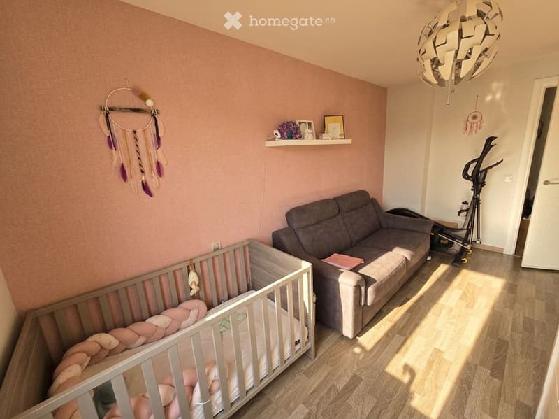 A cozy, well-lit room with a grey sofa, a wooden crib, a shelf with decorative items, and a dreamcatcher hanging on the wall. An exercise bike is positioned near the door.