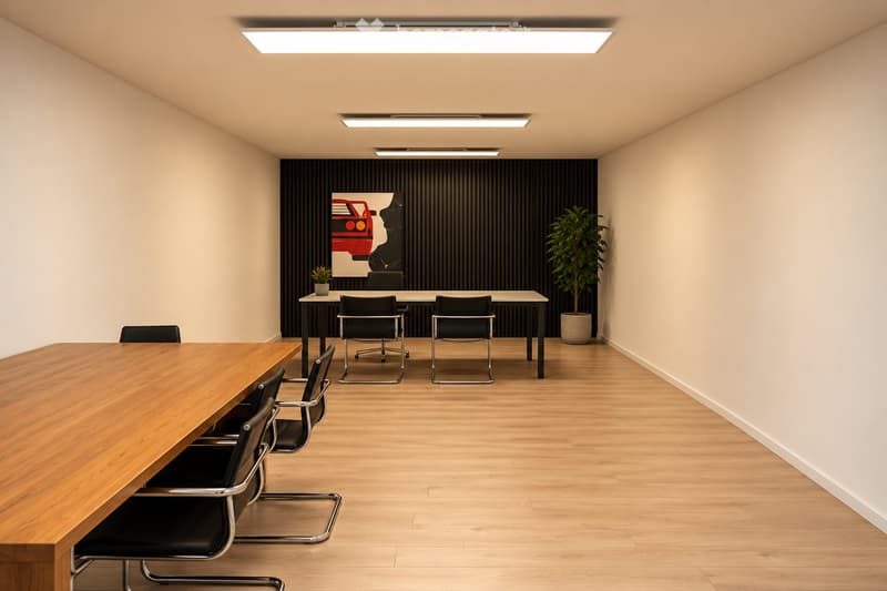 Modern conference room with light wood flooring, two long tables, multiple black chairs, a potted plant, and a large artwork on the wall.