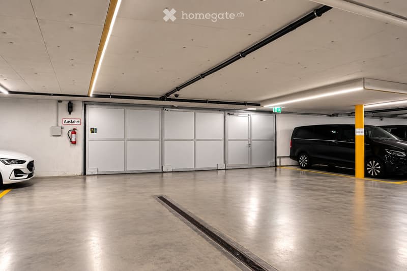 Underground parking area with concrete floors, white walls, and several parked vehicles including a white car and a black van. Features include a large garage door, yellow parking lines, and a fire extinguisher on the wall.