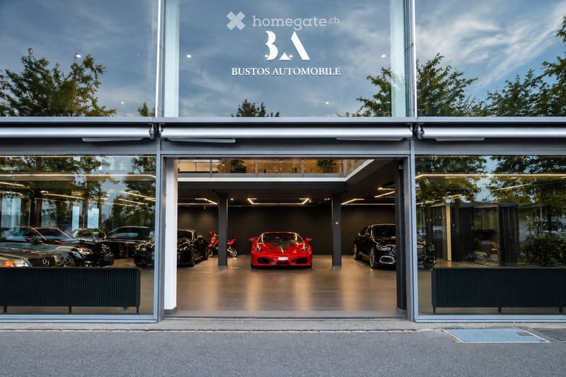 Modern car dealership with large glass facade, displaying several luxury cars inside, including a red Ferrari and several black cars.