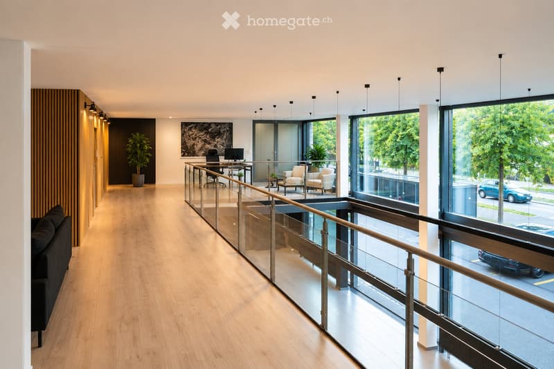 Modern, well-lit corridor with wooden flooring, glass railing, and contemporary furniture including a sofa, chairs, a table, and a computer setup.