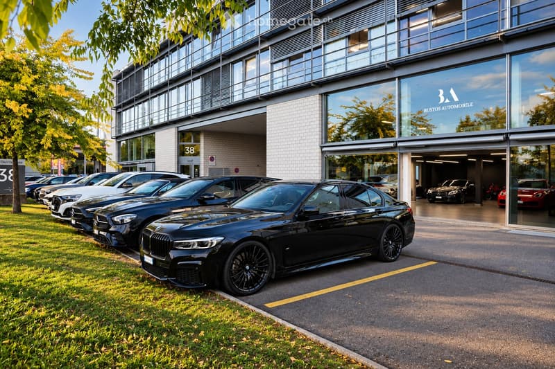 Modern multi-story building with large glass windows and a sleek design, featuring a car showroom with several luxury cars parked outside and inside.