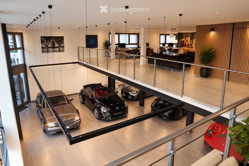 Modern, multi-level showroom with a sleek design, featuring a glass balcony overlooking a lower level with several luxury cars displayed. The upper level includes a seating area with a sofa, chairs, a table, and a large screen, along with office desks and