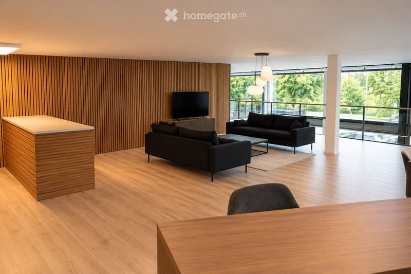 Modern living room with wooden flooring, large windows, and a balcony. Features include a sofa, coffee table, chair, and a screen.