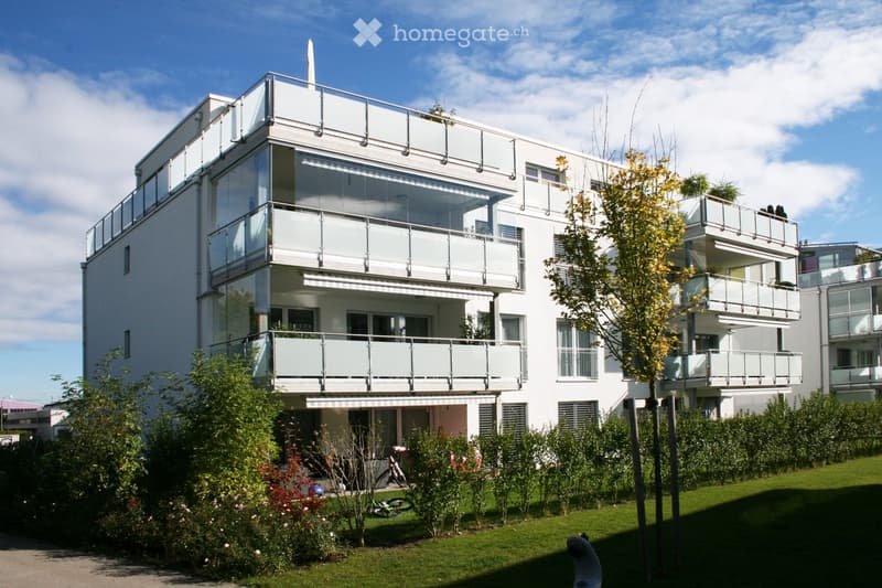 Modern multi-story residential building with white facade, large glass balconies, and surrounding greenery.