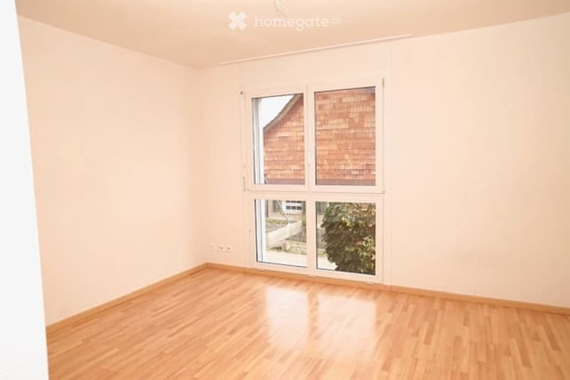 Empty room with light-colored walls and wooden flooring, featuring large windows and a sliding glass door leading to an outdoor area.