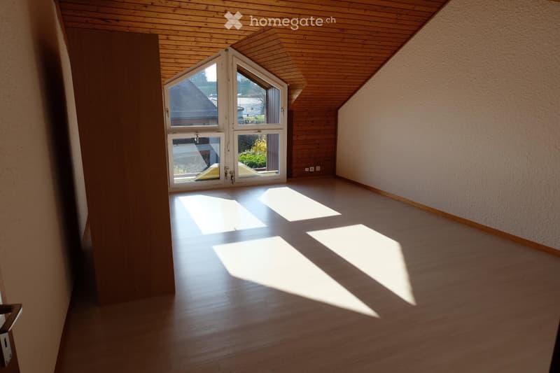 Empty room with wooden flooring, white walls, and a large window with a view of the outside. The ceiling is wooden with a sloped design.