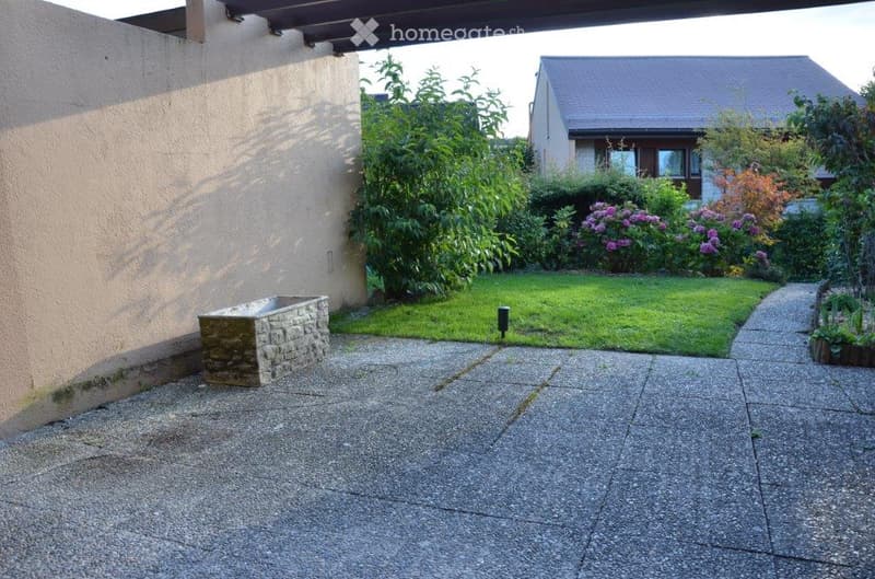 Garden area with a stone planter, well-maintained lawn, and surrounding greenery. The space is paved with concrete tiles and features a pathway leading to a neighboring house.