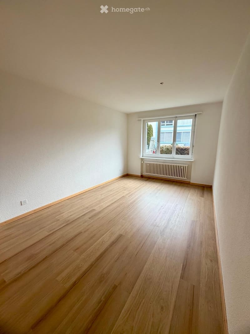 Empty room with light wooden flooring, white walls, and a large window with a radiator below it.