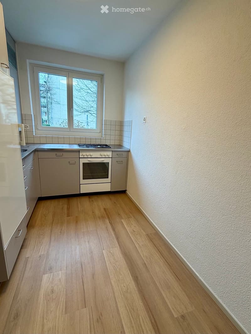 Empty kitchen with wooden flooring, white cabinets, and a window overlooking a building exterior.