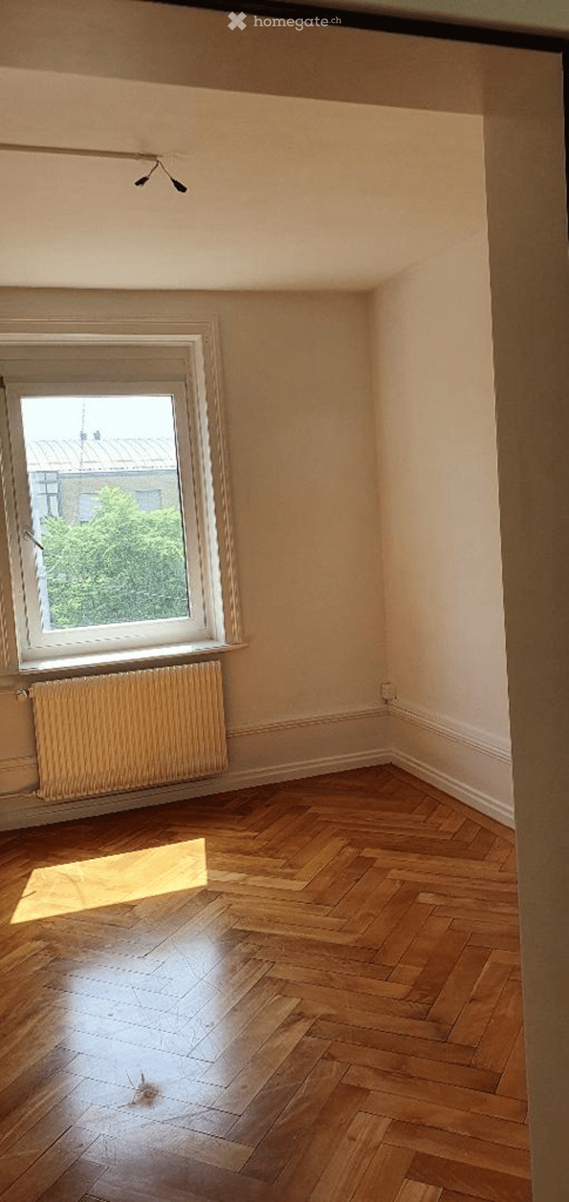Empty room with wooden parquet flooring, white walls, a single window with white frame, radiator below the window, and a small light fixture on the ceiling.