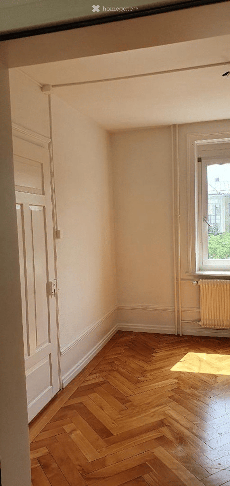 Empty room with wooden herringbone flooring, white walls, and a single window with a radiator below it. The room has a door on one side and is well-lit with natural light.