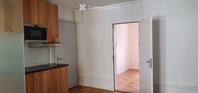 Empty kitchen with wooden cabinets, a microwave, and a sink, leading to an adjacent unfurnished room with wooden flooring.