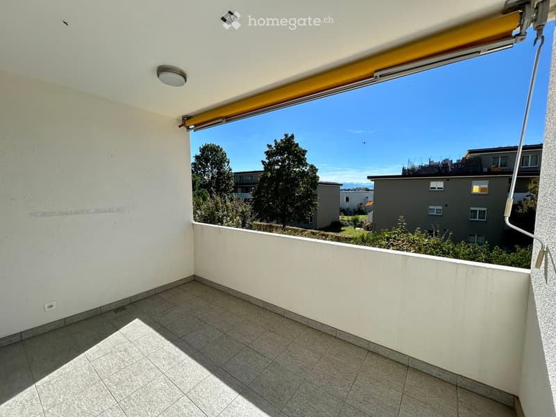 Balcony with tiled floor, white walls, and a retractable awning, overlooking a residential area with trees and buildings.