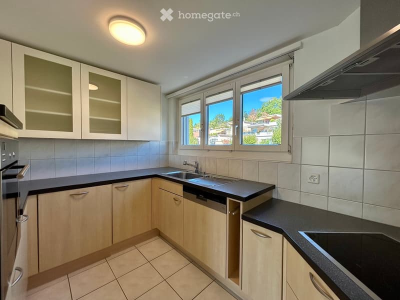 Modern kitchen with white and light wood cabinets, black countertops, stainless steel sink, dishwasher, and range hood. Large windows provide natural light and a view of greenery outside.