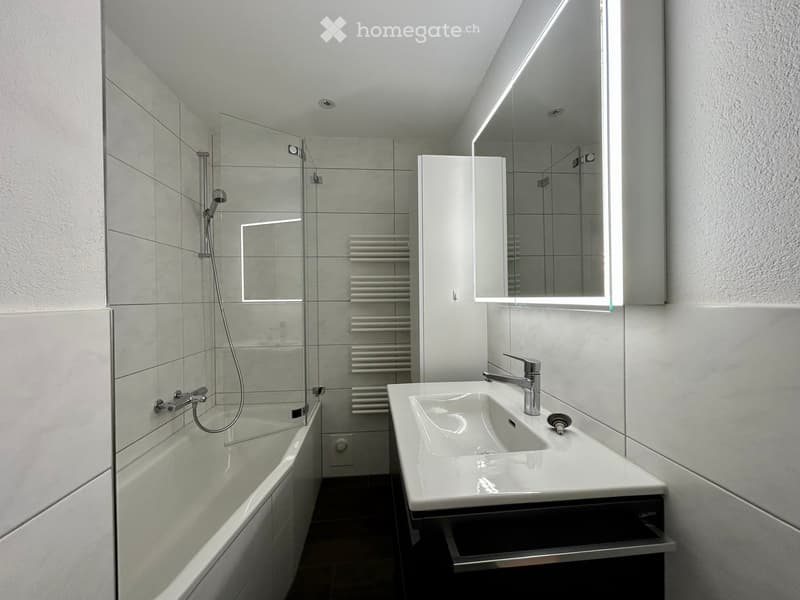 Modern bathroom with a white bathtub, a white sink with a modern faucet, a large illuminated mirror, and a radiator in the corner.