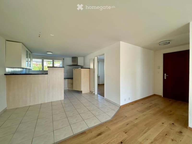 Empty room with wooden flooring and tiled area, featuring a kitchen area with cabinets and a countertop visible in the background.