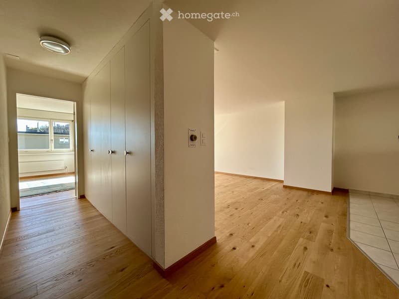 Empty hallway with wooden flooring, white walls, and a closed door leading to another room. Natural light streams in from an adjacent space.